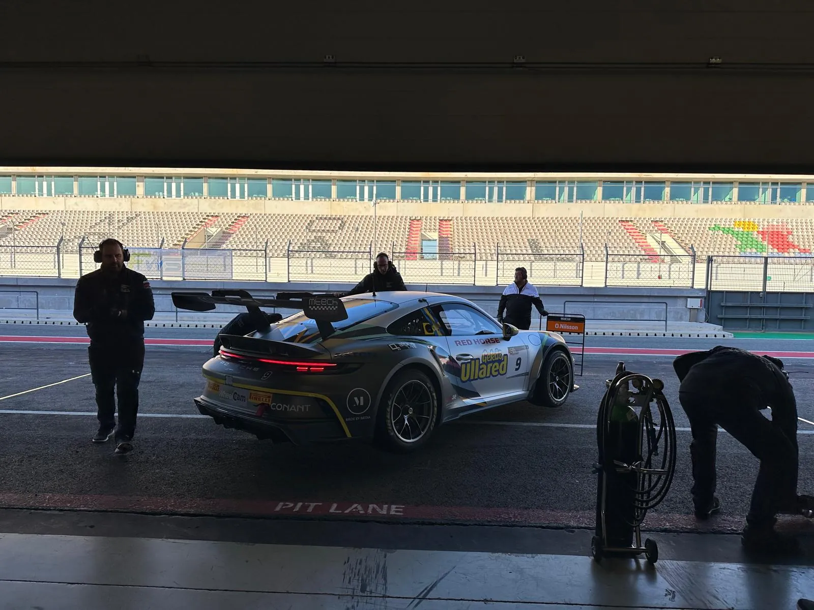 The car in the pit lane at the end of the day, crew around it and empty grandstands behind