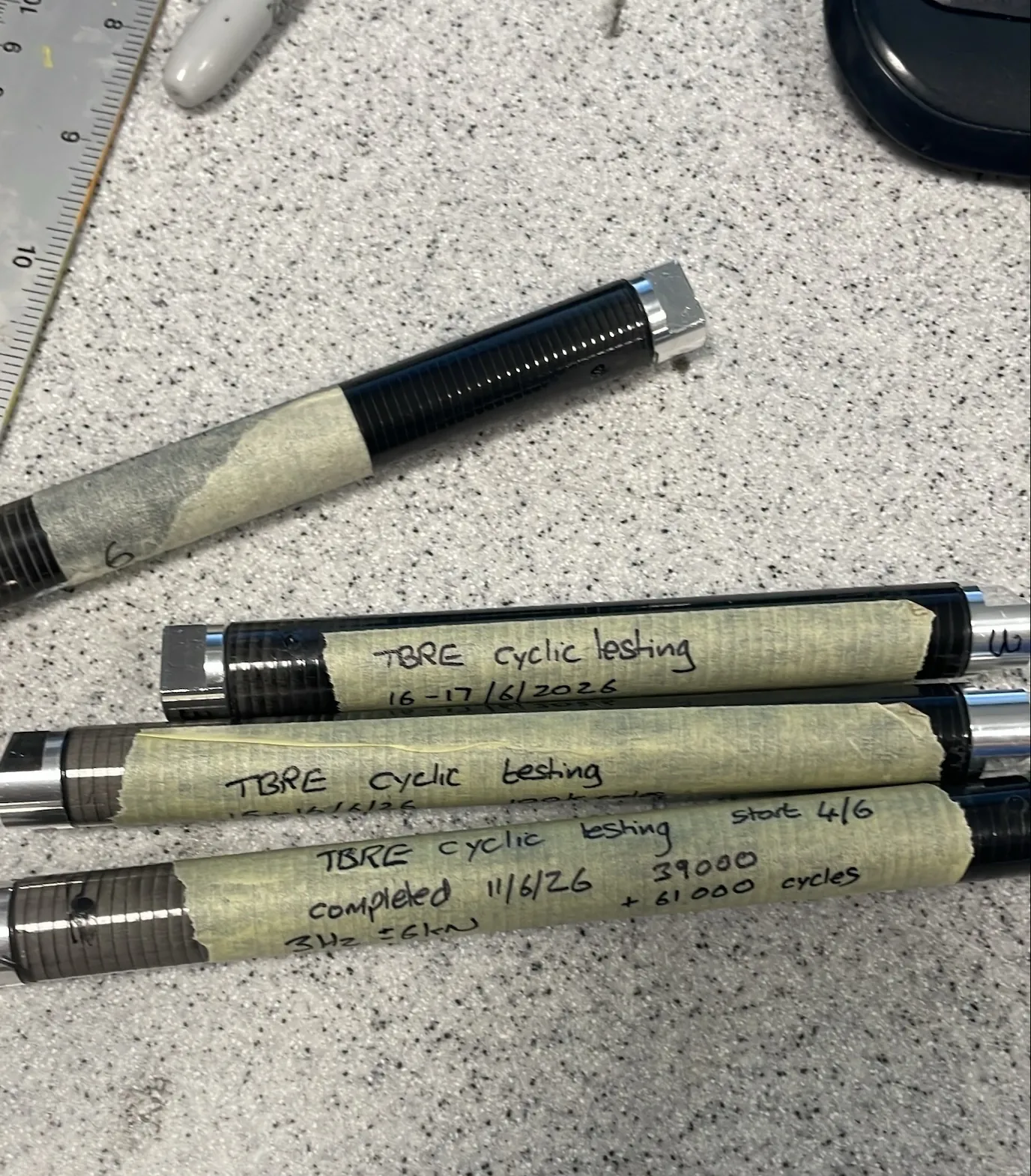 Four representative pushrod members on a bench, each a carbon tube with bonded metallic end fittings, hand-labelled with the dates and cycle counts of their fatigue runs