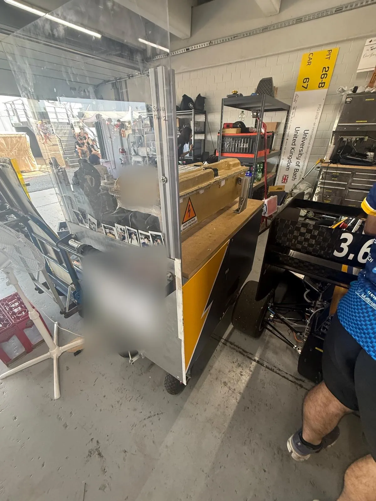 The trolley in the team pit box beside the car, accumulator on the worktop behind the shield, high-voltage warning label on the panel